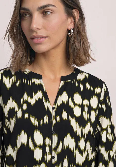 Vrouw met bruin haar in een zwart-wit-geel patroonblouse met V-hals, zijwaarts kijkend.