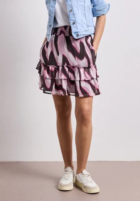 Eine Frau trägt einen gestuften, rosafarbenen und schwarzen gemusterten Rock mit Denimjacke und weißen Sneakers.