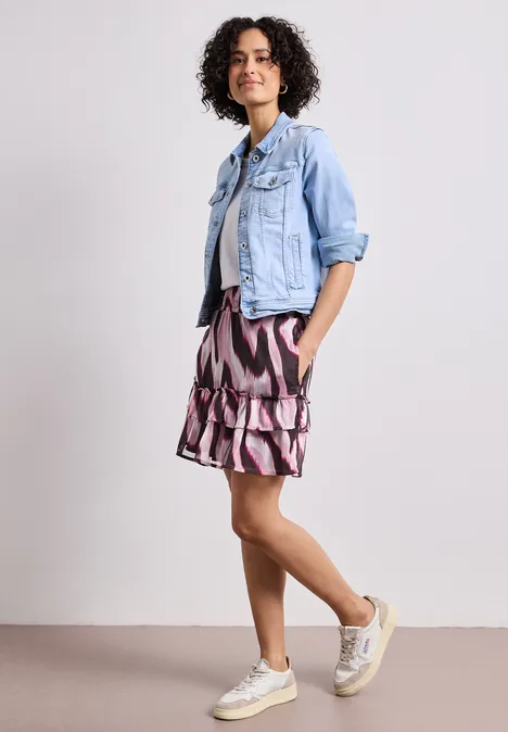 Frau in hellblauer Jeansjacke, gemustertem Rock und Sneakers vor neutralem Hintergrund.