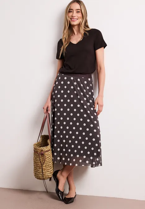 Vrouw in zwarte top en midi-rok met stippen, met strozak en ballerina’s voor een lichte achtergrond.