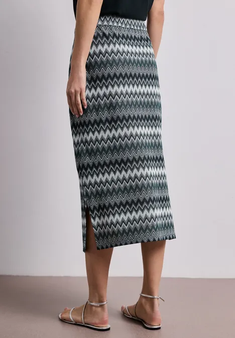 Feminin geschnittener, knielanger Rock mit grauem Zickzackmuster, seitlichem Schlitz und flachen Sandalen.