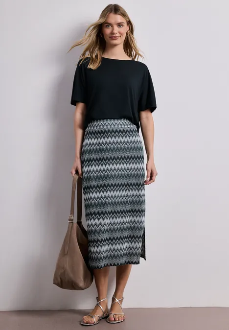 Eine Frau trägt ein schwarzes T-Shirt, einen gemusterten Midirock, Sandalen und hält eine große Tasche.