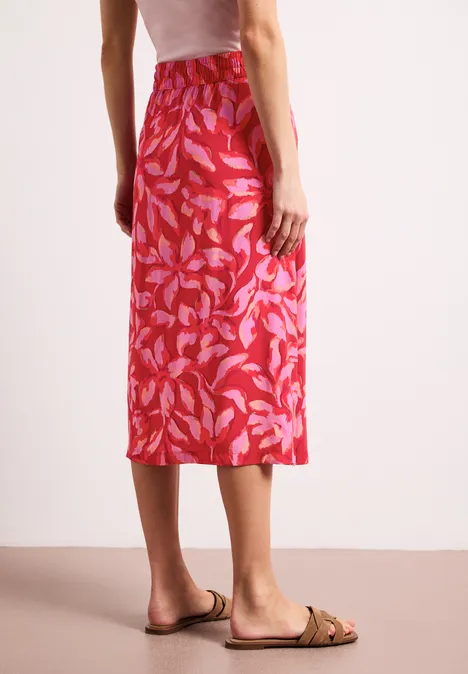 Achteraanzicht van een vrouw in een rode midirok met bladmotief en beige sandalen.