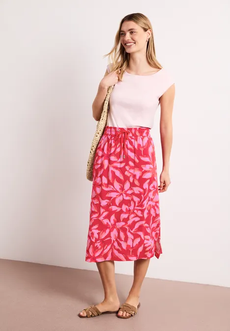 Een vrouw draagt een roze t-shirt, een rode rok met bladprint, sandalen en een strooien tas.