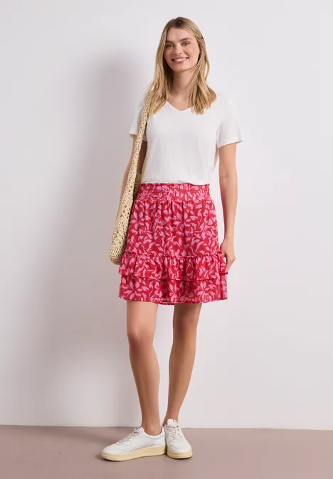 Frau in weißem T-Shirt, rotem gemustertem Rock und weißen Sneakern mit Strohtasche vor hellem Hintergrund.