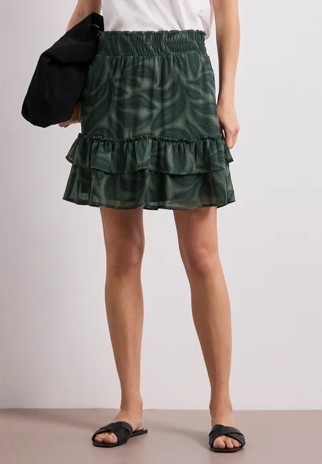 Dunkelgrüner, stufiger Minirock mit Rüschen, weißes Oberteil, schwarze Sandalen und Tasche.
