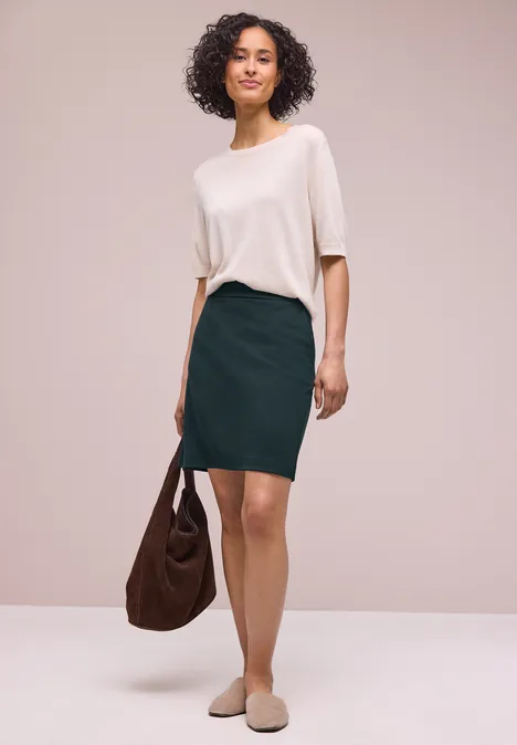 Frau mit lockigem Haar trägt weiße Bluse, grünen Bleistiftrock, beigen Slingback-Pantoffeln und hält eine braune Tasche.