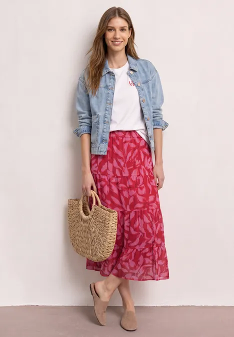 Vrouw in wit shirt, spijkerjasje, rood bloemenrok en rietentas.