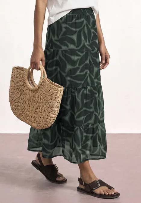 Grüner Midi-Rock mit Blattmuster, braune Sandalen und runde Strohtasche in der Hand.