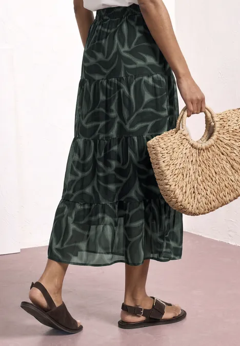 Grüner Maxirock mit Blattmuster, schwarze Sandalen und eine runde Basttasche in der Hand.