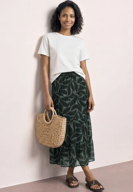 Frau an rosa Wand mit weißem Shirt, grünem Blattmuster-Rock, Strohtasche und Sandalen.