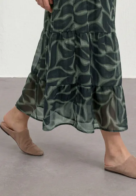 Vrouw draagt groene maxirok met bladprint en transparante volant, met beige mules op grijze vloer.