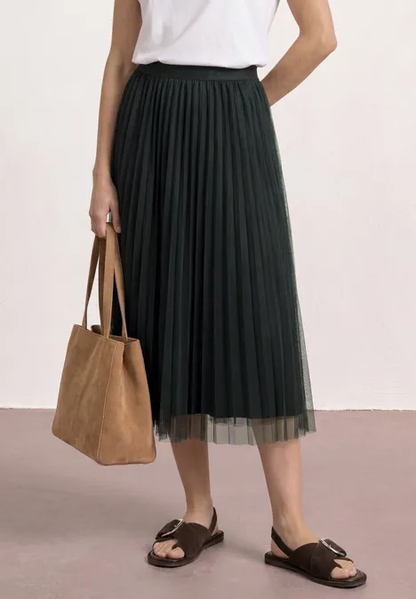 Frau trägt weiße Bluse, schwarzen plissierten Midi-Rock, braune Wildledertasche und schwarze Sandalen.
