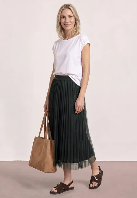 Frau in weißem T-Shirt, schwarzem Plisseerock, schwarzen Sandalen und großer brauner Tasche.
