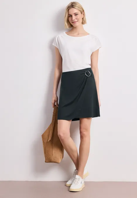 Eine Frau in weißem T-Shirt, dunkelgrünem Wickelrock, weißen Sneakern und brauner Tragetasche.