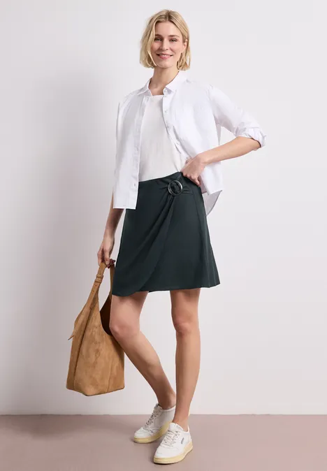 Frau mit weißer Bluse, schwarzem Rock, weißen Sneakers und brauner Tasche vor weißem Hintergrund.