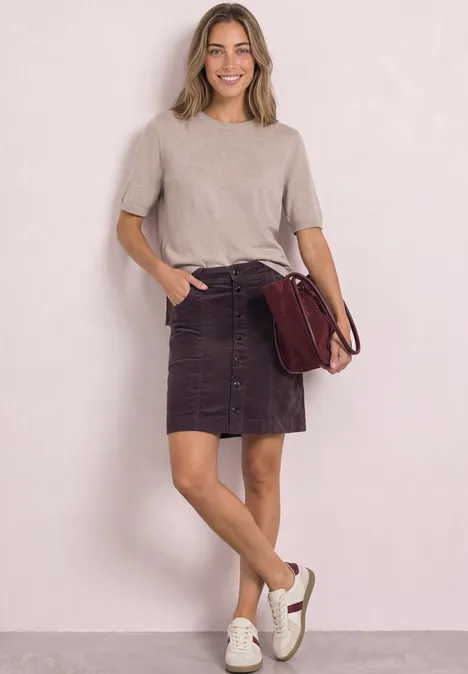 Jeune femme souriante en haut beige, jupe violette à boutons, baskets blanches et sac bordeaux.
