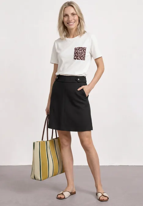 Femme en t-shirt blanc à poche, jupe noire, sandales et grand sac rayé.
