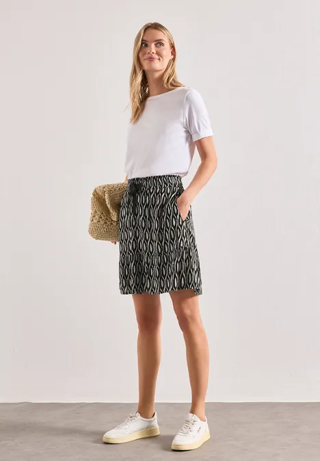 Jupe au genou à imprimé et à volants clay sand Femme en t-shirt blanc, jupe à motifs et baskets blanches, tenant un sac en paille.