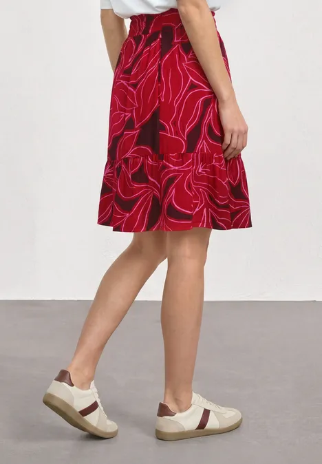 Person trägt roten, floralen Rock und weiße Sneaker, blickt seitlich.