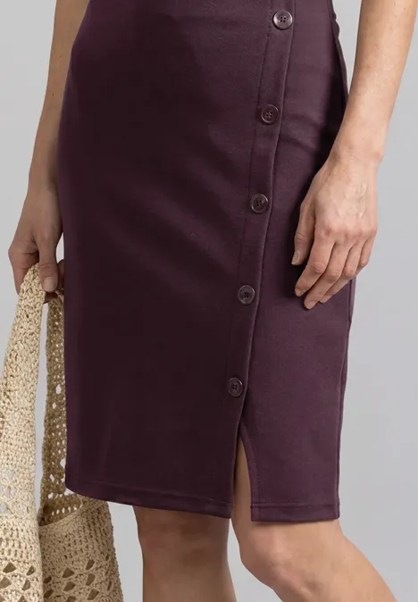 Jupe crayon aubergine mi-longue avec rangée de boutons latéraux et fente, accompagnée d’un sac filet en crochet.
