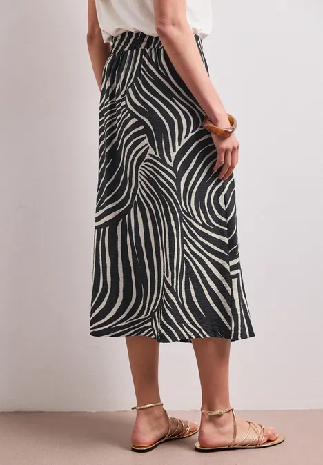 Frau in Midirock mit Schwarz-Weiß-Zebra-Muster und beigen Sandalen vor schlichter Wand.