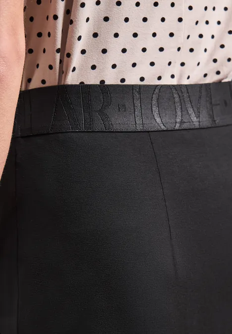 Frau trägt weiße Bluse mit schwarzen Punkten und schwarze Hose mit geprägtem Schriftzug am Bund.