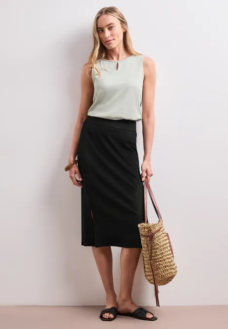 Vrouw in wit mouwloos topje, zwarte midirok met split, zwarte sandalen en rieten tas.