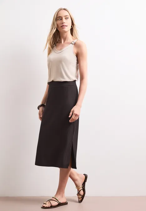 Frau mit weißem Top, schwarzem Midirock und Sandalen vor weißem Hintergrund.