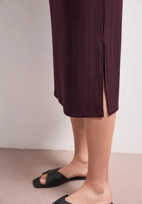 Schwarze Sandalen mit geknotetem Riemen, burgunderrotes Midikleid, Rippenoptik mit Seitenschlitz vor beigem Hintergrund.