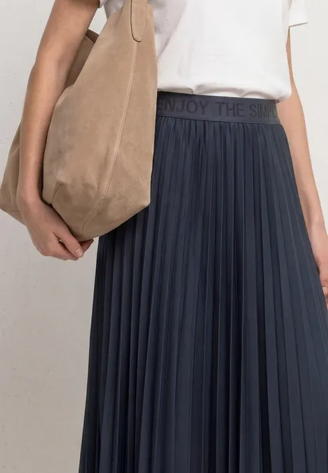 Spódnica midi o plisowanej strukturze darkest blue Kobieta w białym t-shircie, granatowej plisowanej spódnicy i beżowej torbie na ramię.