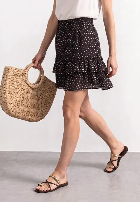 Kniehoge chiffonrok met print black coffee Eine Frau in weißem Top, schwarzem, gepunktetem Rock, mit Basttasche und schwarzen Riemchensandalen.