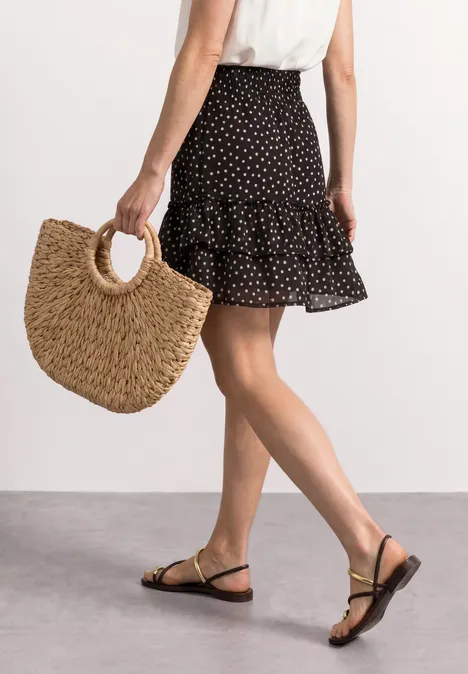 Kniehoge chiffonrok met print black coffee Frau in Tupfenrock, weißem Top und schwarzen Sandalen mit goldenen Details trägt eine geflochtene Strohtasche