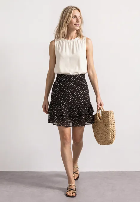 Kniehoge chiffonrok met print black coffee Frau in ärmelloser weißer Bluse und schwarzem, gepunktetem Rock mit Stroh-Tasche und Sandalen.