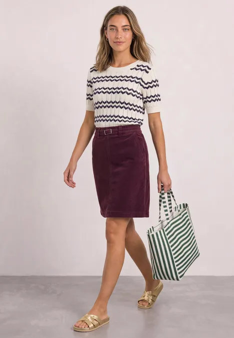 Vrouw in T-shirt met patroon, paarse rok, gouden sandalen en groen-witte draagtas.