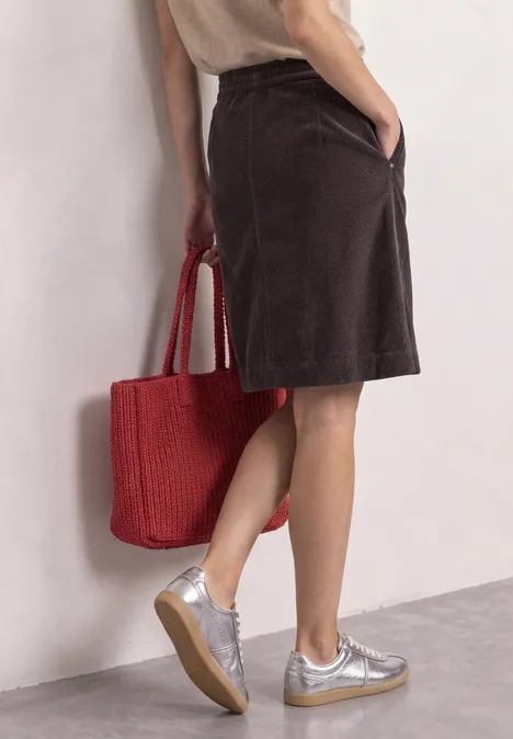 Vrouw met bruine midirok, zilveren sneakers en een rood gevlochten katoenen tas.