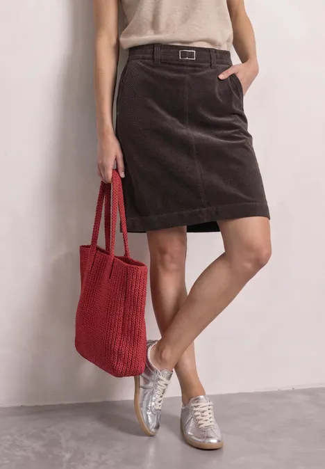 Persoon met bruine corduroyrok, beige top, witte sneakers en rode stoffen tas voor een lichte muur.