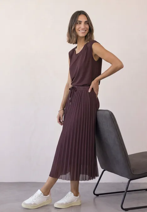 Midirok van mesh met plissé-structuur rustic rouge Eine Frau in bordeauxrotem ärmellosen Oberteil und Plisseerock, weiße Sneaker, steht neben einem Stuhl.