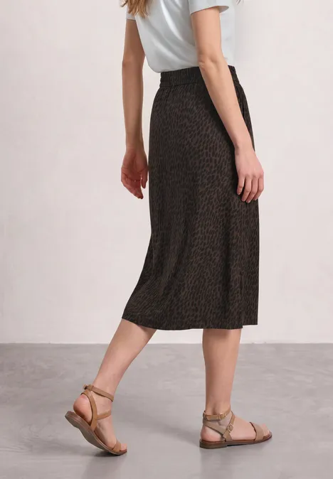 Jupe mi-longue à taille élastiquée et fentes molten earth Vue latérale d’une femme en haut blanc, jupe midi noire à motif discret et sandales fines plates.
