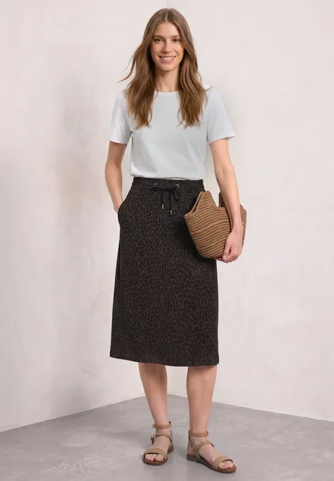 Jupe mi-longue à taille élastiquée et fentes molten earth Femme en t-shirt blanc, jupe noire midi à motifs discrets, sandales et sac en osier sur fond clair.
