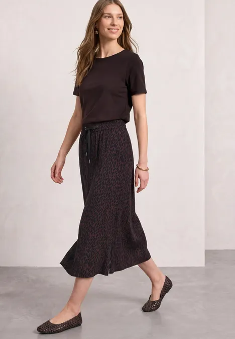 Frau in schwarzem T-Shirt und dunklem, gepunktetem Midirock mit Kordelzug, dazu schwarze Ballerinas, stehend.
