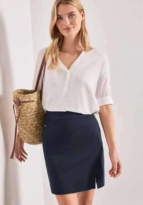 Une femme porte une chemise blanche, une jupe crayon bleu foncé et un grand sac en paille.