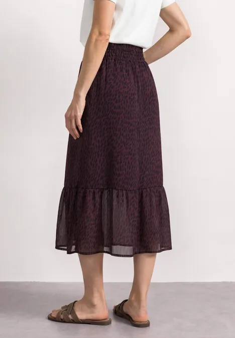 Eine Frau trägt weißes Shirt und dunklen gemusterten Midirock mit Rüschen, flache Sandalen.