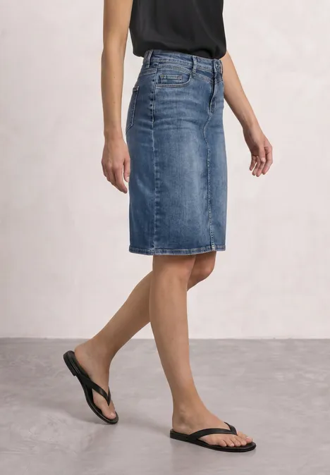 Frau in knielangem Jeansrock, schwarzem Top und Flip-Flops vor schlichter Wand.
