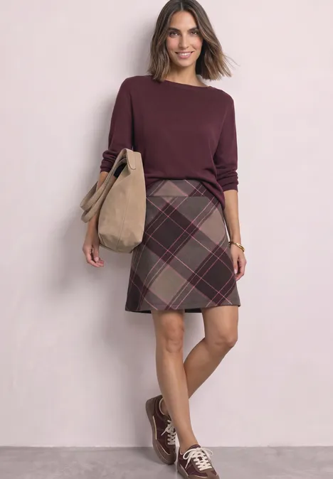 Femme en pull bordeaux, jupe à carreaux et sac bandoulière beige.