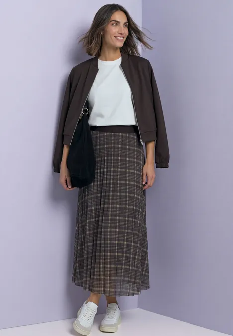 Femme en veste marron, top blanc, jupe longue à carreaux et baskets blanches sur fond violet clair.