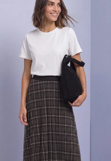 Une femme souriante devant un fond violet porte un t-shirt blanc, une jupe à carreaux foncés et tient un sac noir.
