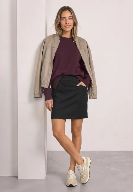 Vrouw in zwarte minirok, bordeaux trui, beige jasje en witte sneakers voor een lichte achtergrond.