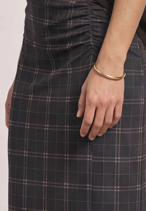 Frau in schwarzem kariertem Kleid mit gerafftem Detail, Arm mit goldenem Armreif sichtbar.