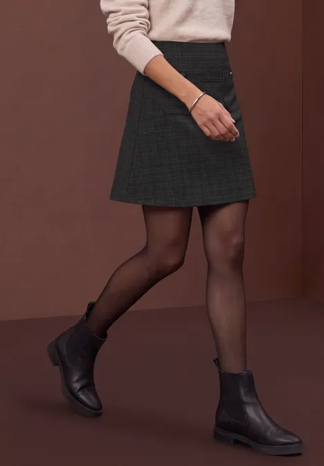 Une personne en jupe courte, collants noirs et bottines noires marche devant un fond brun.